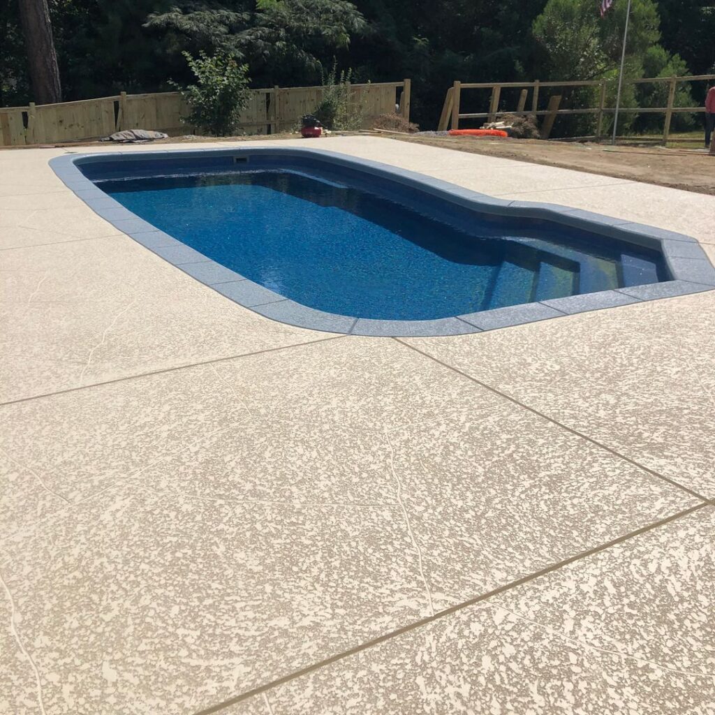A high-angle view of a dark blue in-ground swimming pool with built-in steps, surrounded by a light cream-colored decorative concrete deck. The deck features a textured, speckled finish with thin, tan-colored score lines forming large rectangular sections. The pool is bordered by a grey coping.