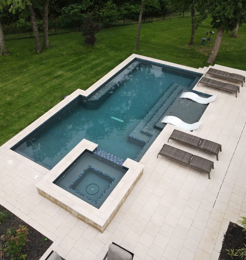 A high-angle view of a modern rectangular swimming pool and integrated square hot tub set within a light-colored stone patio. The pool features dark teal water, a tanning ledge with two white wavy lounge chairs, and several grey wicker sun loungers arranged on the deck.