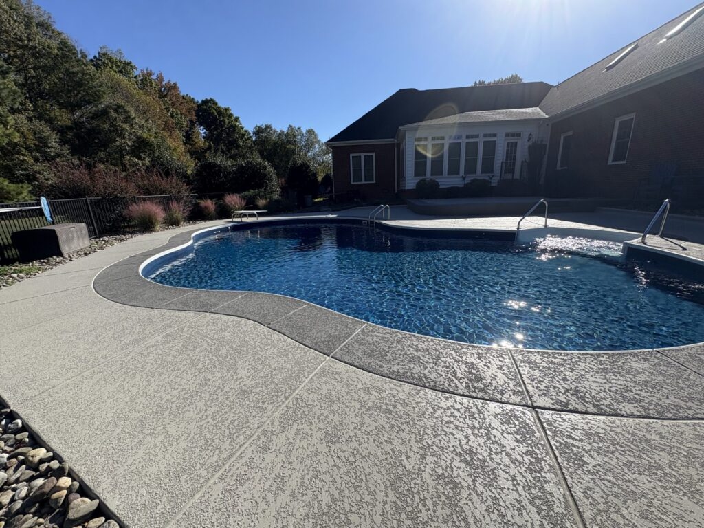 A wide-angle view of a kidney-shaped swimming pool with a deep blue liner and a built-in tanning ledge with white metal handrails. The pool is surrounded by a light tan textured concrete deck featuring a dark gray border and visible expansion joints.