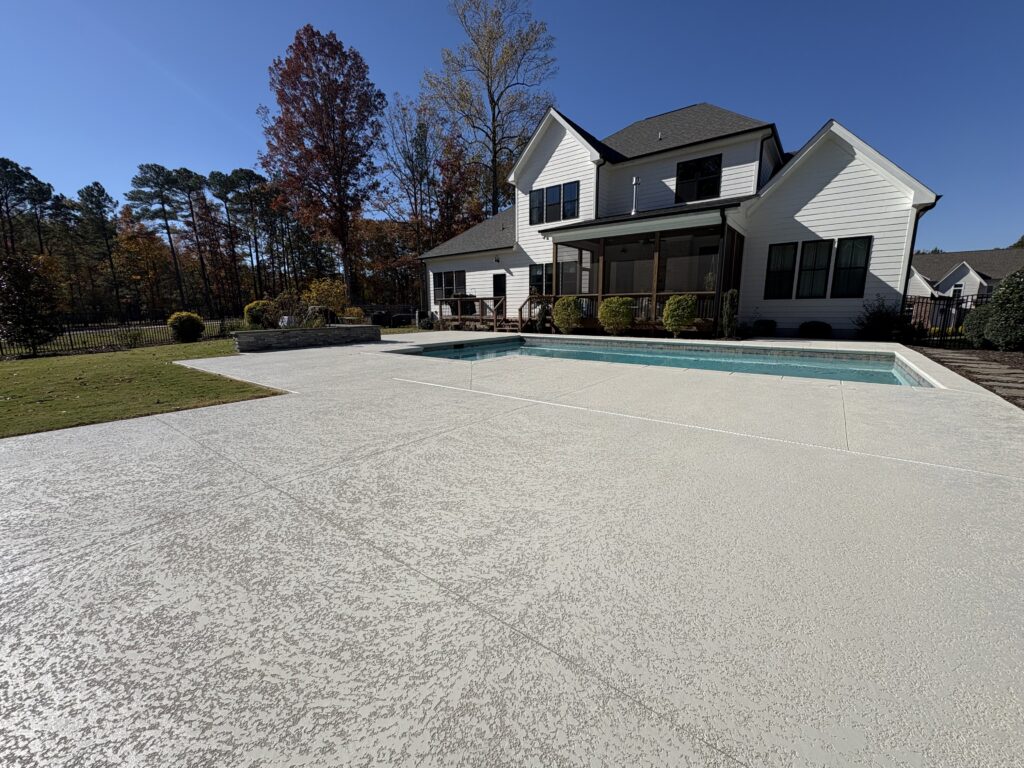 A wide-view of a modern residential backyard featuring a large decorative concrete pool deck with a light grey, textured "knockdown" finish. The expansive patio surrounds a rectangular in-ground swimming pool with light blue water.
