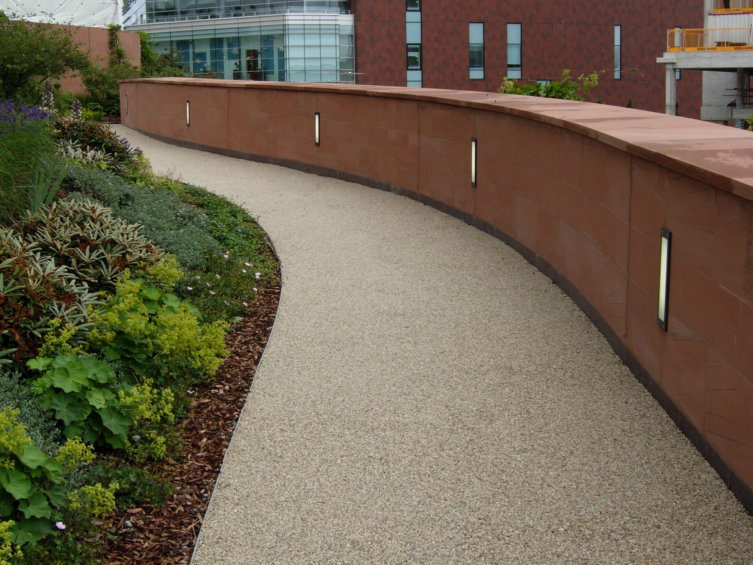 A ground-level view of a winding, beige resin-bound gravel pathway bordered by a low, curved masonry wall in a terracotta shade. The wall features integrated rectangular vertical lights, and the path is lined with lush green shrubs and garden mulch.