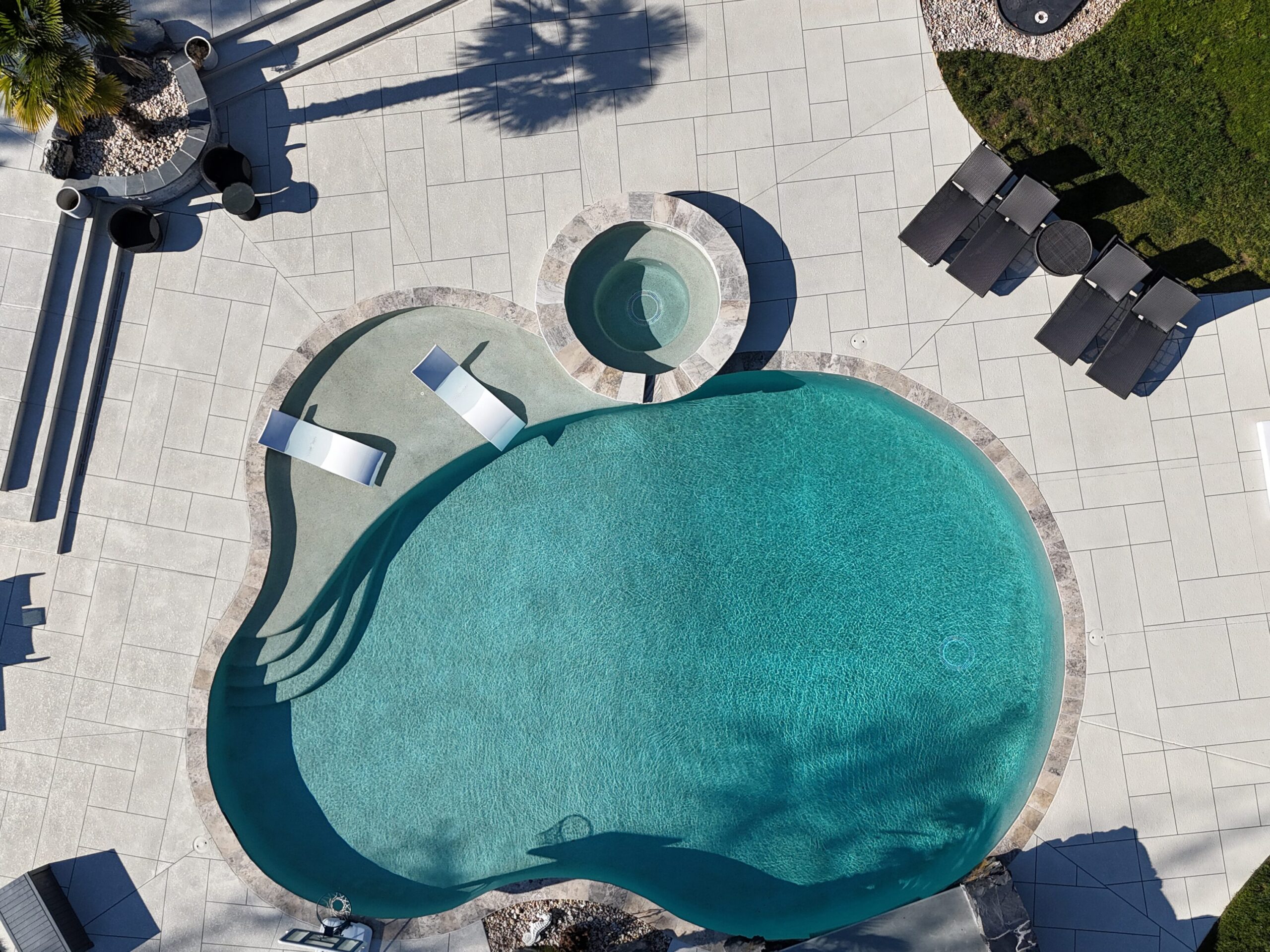 A top-down view of a modern, freeform swimming pool with turquoise water. The pool features a built-in sun shelf with two white lounge chairs partially submerged and a circular, stone-tiled hot tub integrated into the top edge. The surrounding patio is made of large, light-gray rectangular pavers.