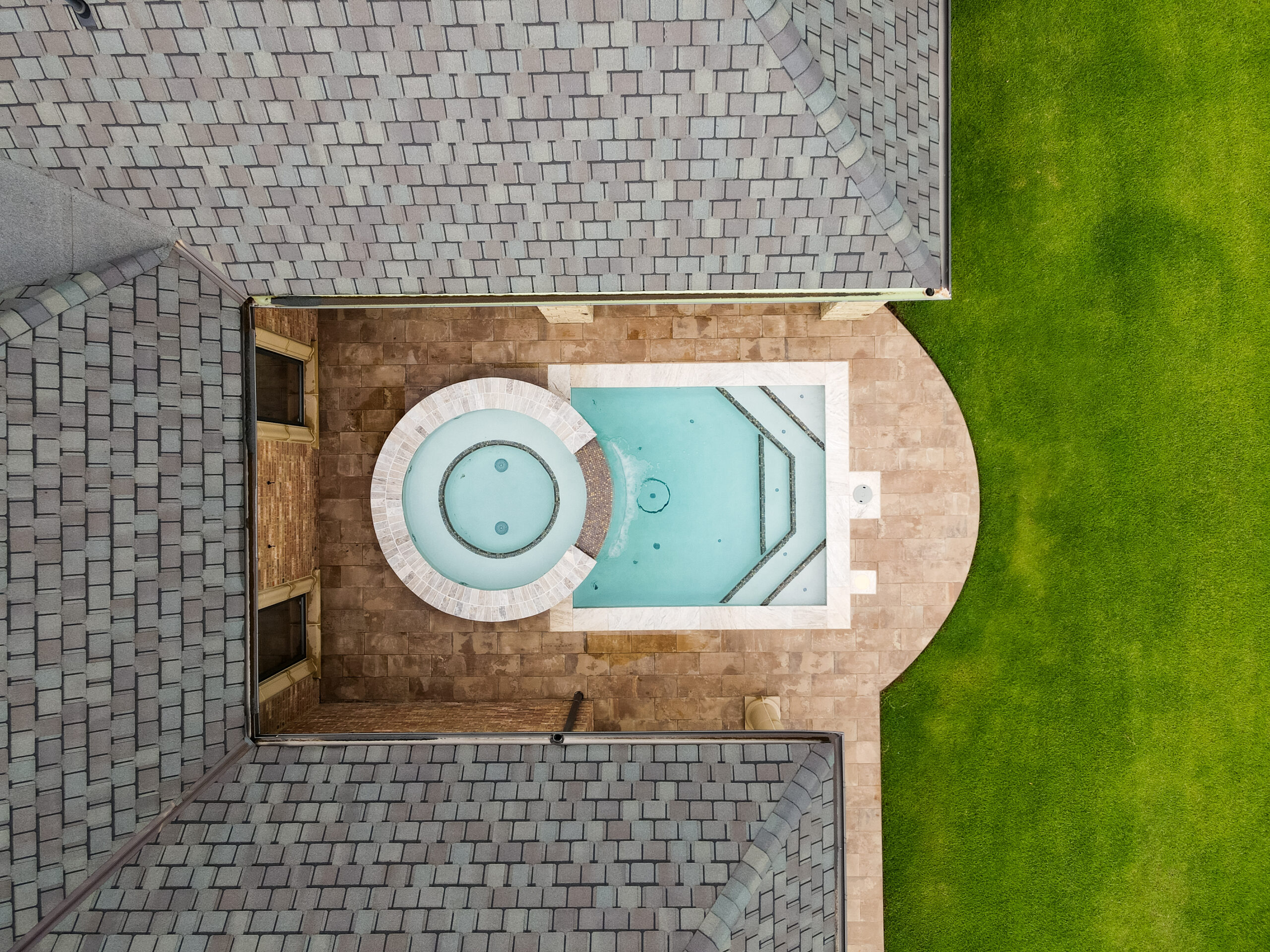 A top-down view of a modern luxury backyard featuring a geometric swimming pool and an integrated circular hot tub. The pool area is nestled into a stone patio within the corner of a house's grey shingled roofline.
