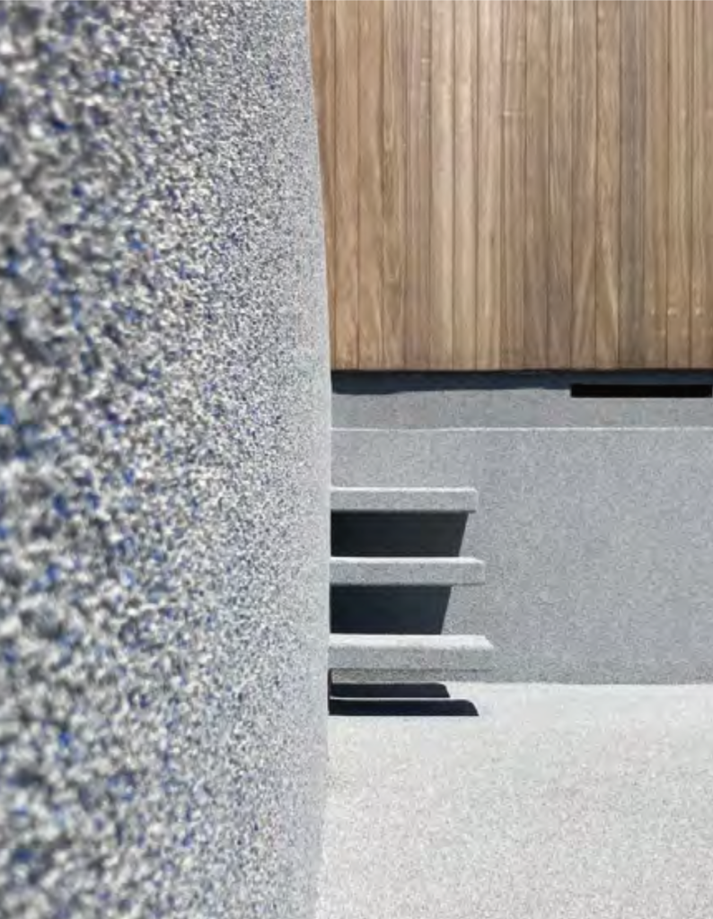 A close-up shot of architectural elements featuring a curved, textured grey stone wall in the foreground. In the background, three cantilevered stone steps protrude from a matching grey wall, casting sharp shadows. Above the stairs, a section of vertical wood paneling provides a warm, natural contrast to the cool-toned, pebbled masonry.