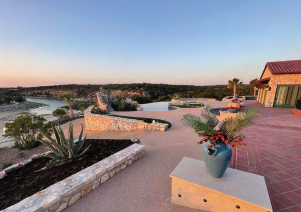 Wide-angle view of a Mediterranean-style stone patio overlooking a river at sunset, featuring an infinity pool, agave plants, and a large blue vase with red flowers in the foreground.