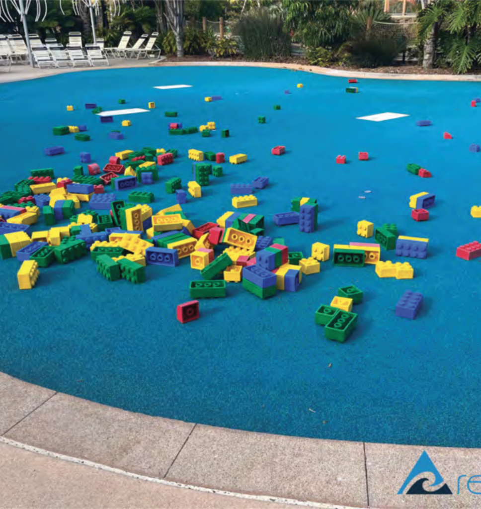 A wide shot of a blue, padded outdoor play surface scattered with numerous large, colorful plastic building blocks in primary colors like red, yellow, green, and blue.