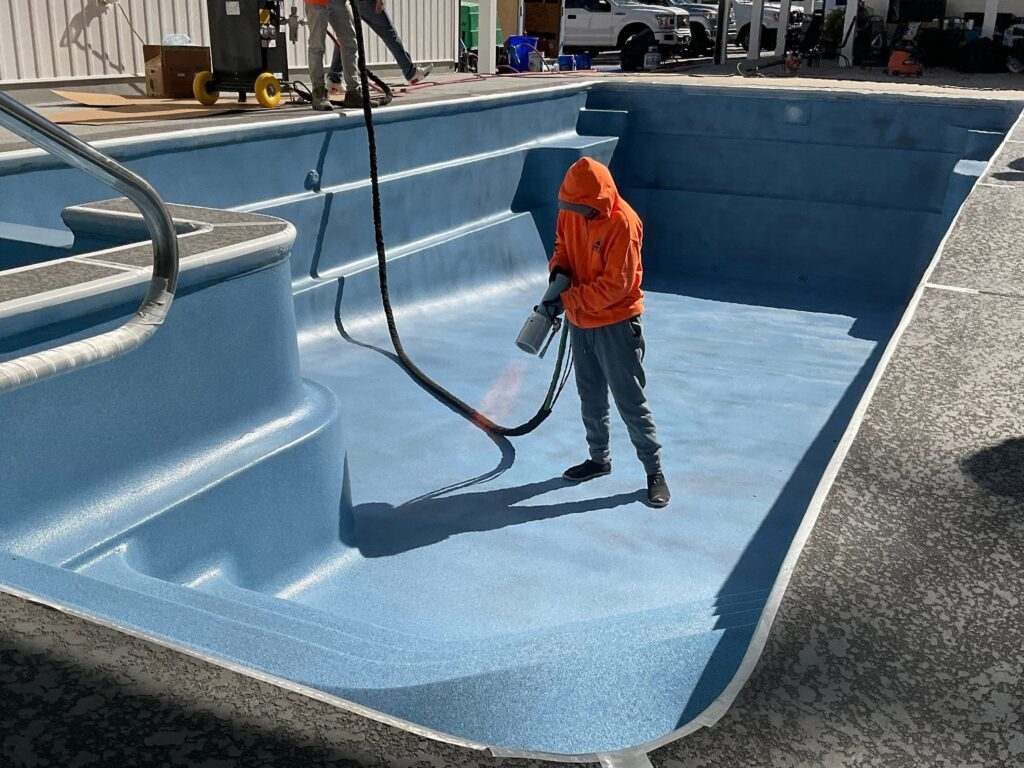 A professional technician in an orange hoodie using an industrial sprayer to apply a blue slip-resistant coating to the floor of an empty in-ground swimming pool. The pool features built-in steps and is surrounded by a textured, gray stamped concrete deck.