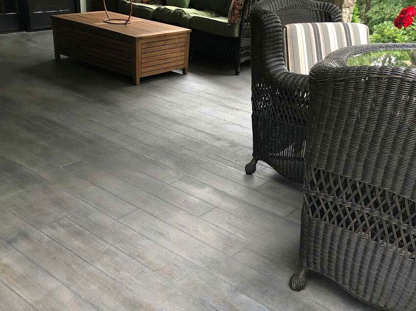 A close-up view of a covered patio featuring grey stamped concrete textured to look like weathered wooden planks. The space is furnished with dark brown wicker armchairs with striped cushions and a wooden slat coffee table.