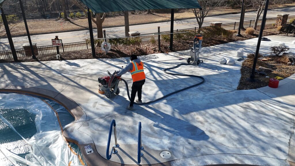 An overhead shot shows a Sundek of NC worker in a bright orange safety vest operating a walk-behind concrete floor grinder on a pool deck. The pool area is protected with plastic sheeting, and the concrete surface shows visible signs of surface preparation and grinding.