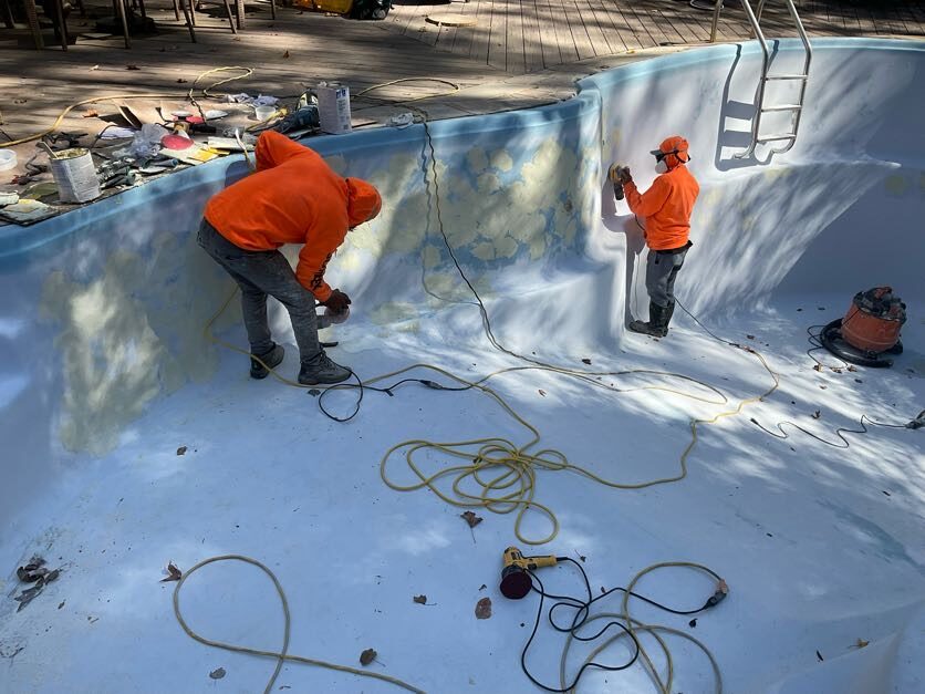wo professional pool technicians in safety gear and orange hoodies use hand grinders to prepare and smooth the surface of a fiberglass pool for resurfacing.